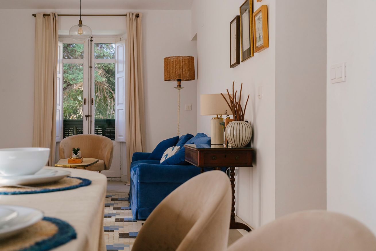 Une pièce avec une table à manger dressée pour deux personnes, un canapé bleu, une lampe et une vue sur l’extérieur, visible à travers une porte.