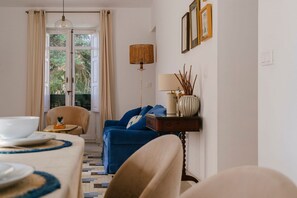 Ein Zimmer mit Esstisch für zwei Personen, einem blauen Sofa, einer Lampe und Blick durch eine Tür.