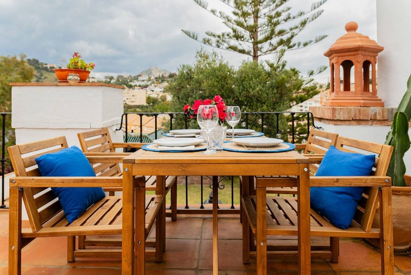 Ein Holztisch für zwei Personen gedeckt, mit einer Vase roter Blumen, zwei Weingläsern und einem Topfpflanzen auf einer Terrasse mit Landschaftsblick.