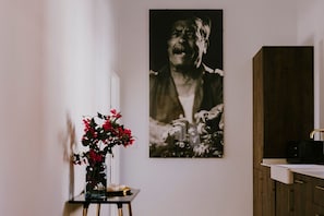 Ein Zimmer mit weißer Wand, einem Schwarzweißfoto, einem kleinen Tisch mit einer Vase roter Blumen und einem Holzschrank mit Waschbecken.