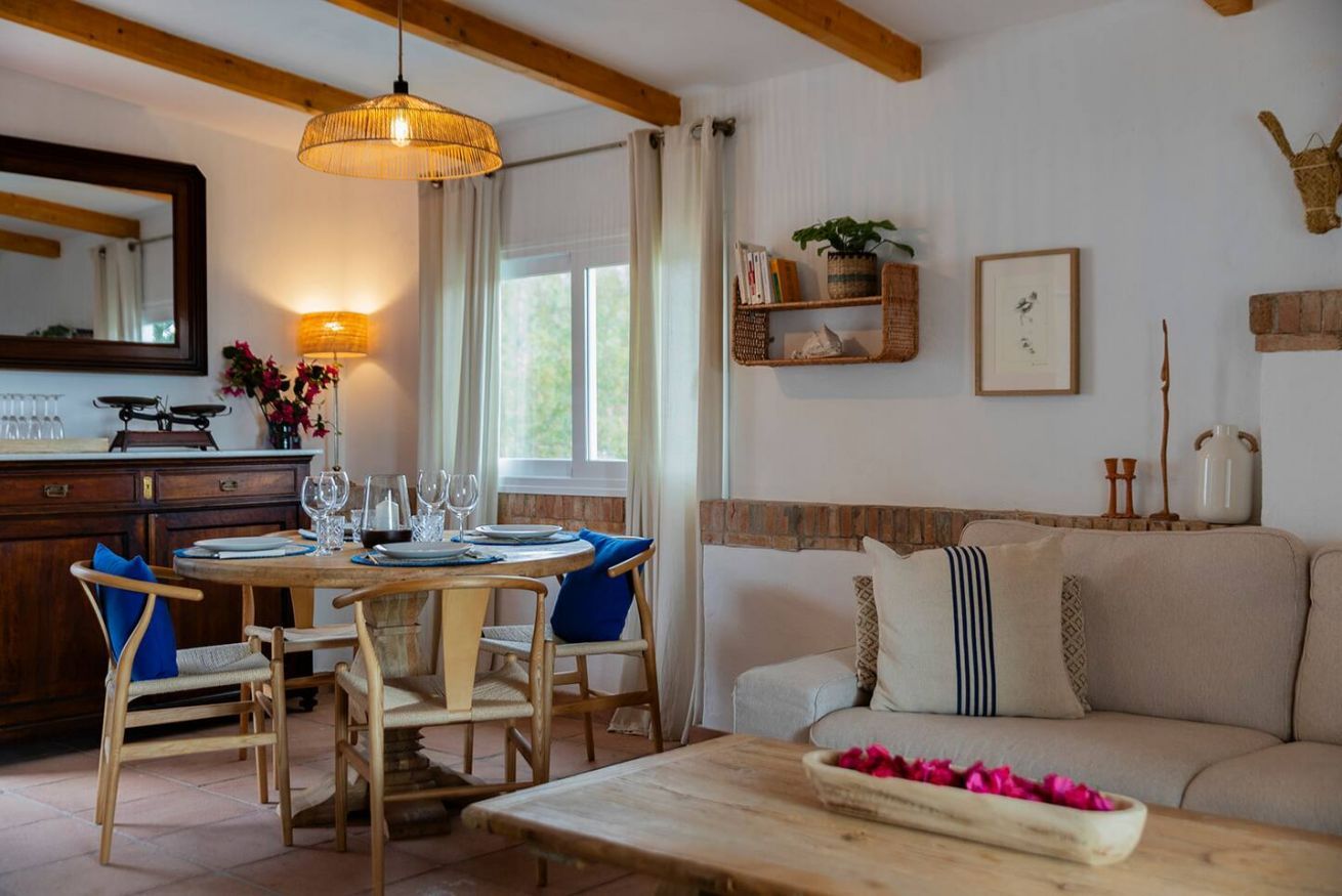 A dining area with a wooden table and chairs, a hanging light fixture, a sofa, and a wall-mounted shelf with decorative items.