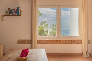 A room with a view of the sea, a wooden table with a flower vase, and a bookshelf with books and decorative items. - Hacienda Clavero Eight Ocean View Apartment (Málaga)