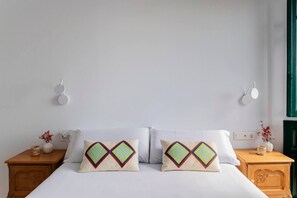 Ein Schlafzimmer mit einem Bett, zwei Kissen mit geometrischen Mustern, einem Nachttisch aus Holz, einer Wandleuchte und einer Vase mit getrockneten Blumen.