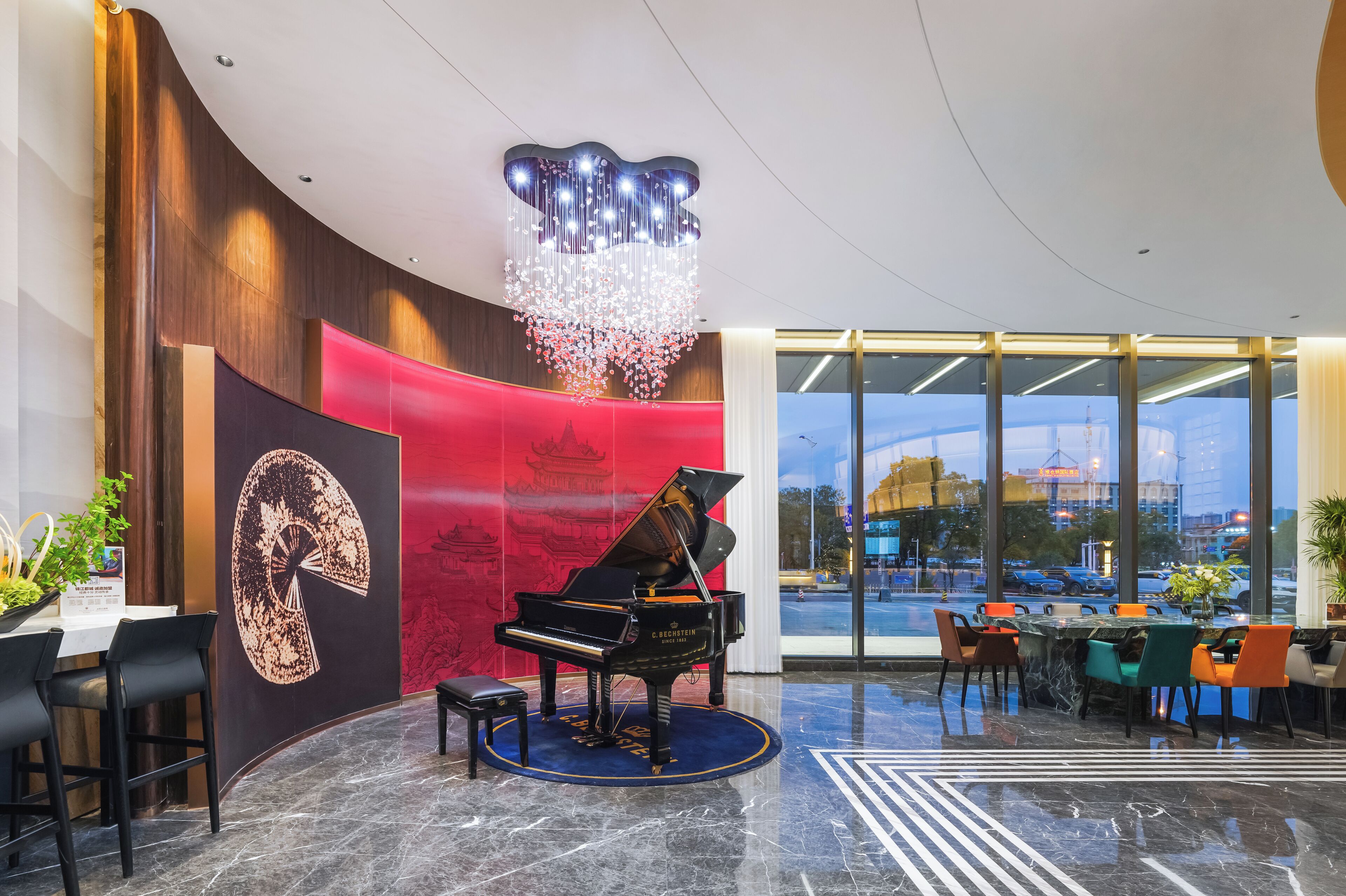 A grand piano in a lobby with a large chandelier, a red wall with a traditional Chinese painting, and a view of a cityscape through large windows.