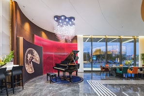 A grand piano in a lobby with a large chandelier, a red wall with a traditional Chinese painting, and a view of a cityscape through large windows. - Metropolo Hotel - Changsha Taskin Plaza (Changsha)