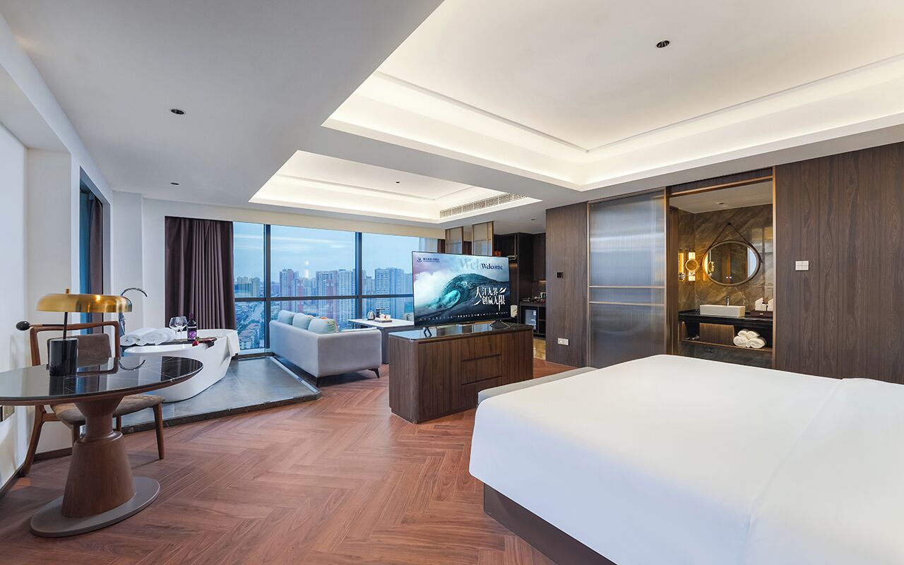 A modern hotel room with a large bed, a dining table, a sofa, a TV, and a city view.