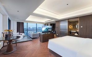 Ein modernes Hotelzimmer mit einem großen Bett, einem Esstisch, einer Sitzecke und einem Fernseher, mit Blick auf die Stadt.