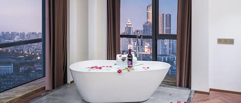 A modern bathroom with a freestanding bathtub, a city view through large windows, and rose petals scattered on the floor.