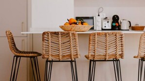 Una cucina con un piano di lavoro bianco, tre sedie in vimini con gambe in metallo nero e una ciotola di arance sul bancone.