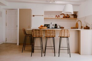 Una cocina con encimera blanca, tres taburetes de mimbre y un lámpara colgante, además de una puerta que da a otra habitación.