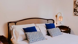 Un letto con biancheria bianca e due cuscini decorati, un comodino con una lampada, e un'opera d'arte appesa al muro.