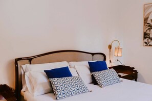 Ein Bett mit weißer Bettwäsche und zwei gemusterten Kissen, ein Nachttisch mit Lampe sowie ein an der Wand hängendes Kunstwerk.
