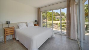 Et hotelværelse med en stor seng, et natbord, en lampe og udsigt til en balkon med grønne planter.