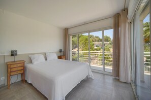 Ein Hotelzimmer mit einem großen Bett, einem Nachttisch, einer Lampe und Blick auf einen Balkon mit Grünpflanzen.