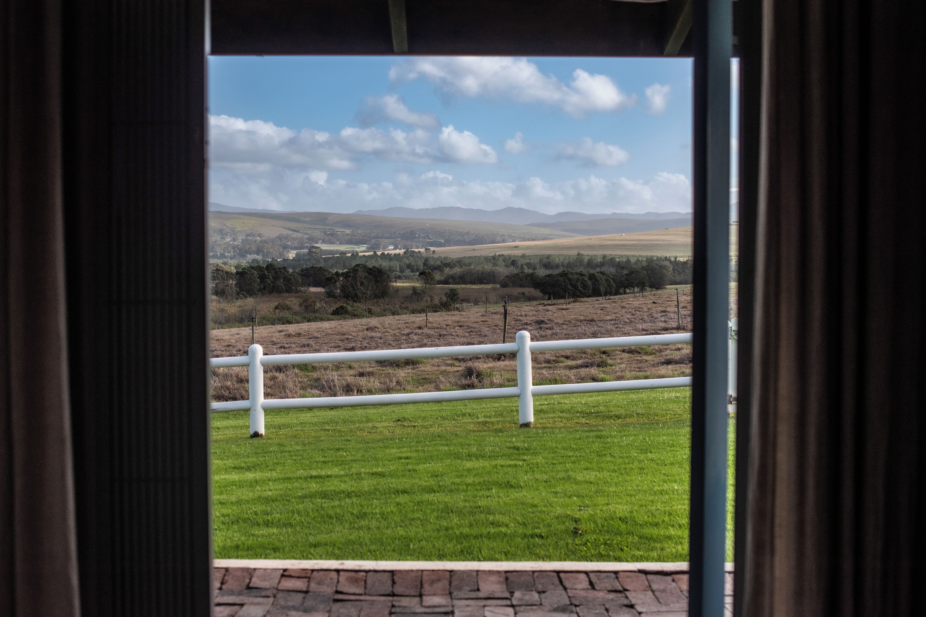 A view through a window with a white railing, a grassy field, and distant hills.