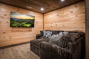 A cozy room with wooden walls, a large TV mounted on the wall, a cushioned sofa with a blanket, and a small wooden side table. - Putts & Pines | King Suites with Putt-Putt, Pool, Golden Tee, Buck Hunter (Sevierville)