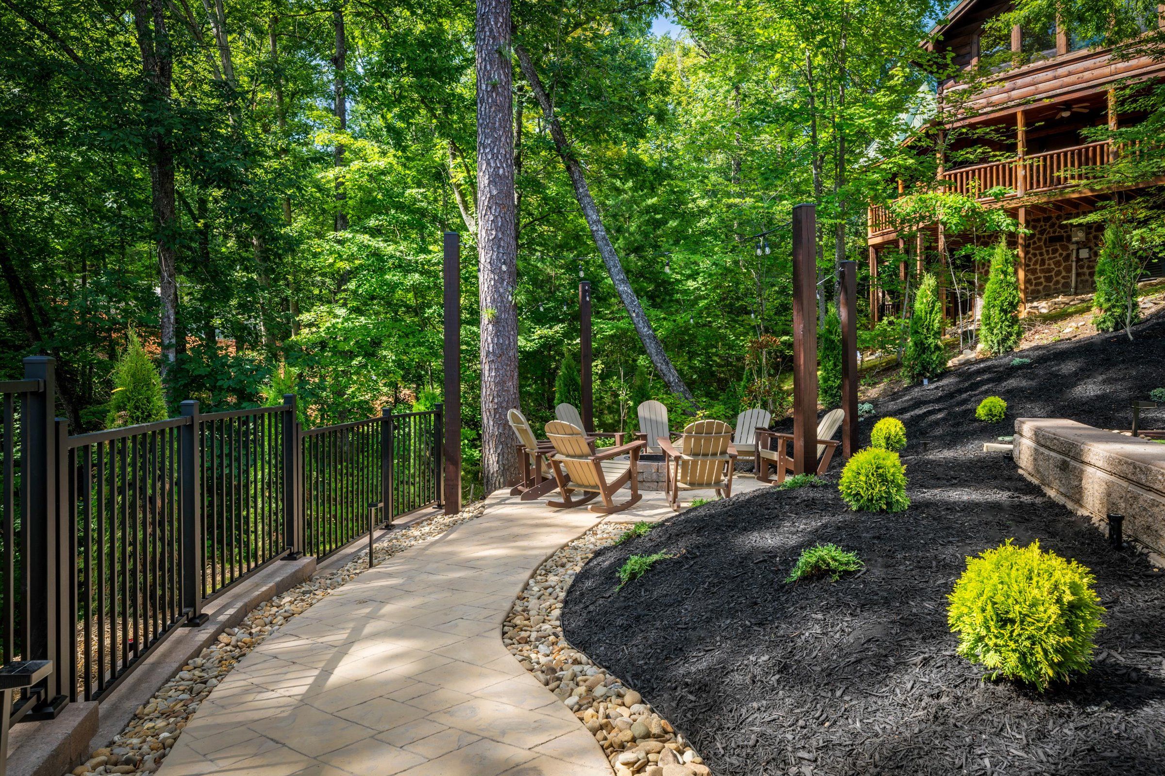 A paved pathway leading to a seating area with Adirondack chairs, surrounded by a wooden fence and a stone wall.
