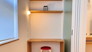 A corner room with a wooden desk, a red stool, a shelf with a picture, and a wall-mounted light.
