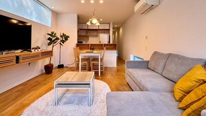 A modern living room with a flat-screen TV, a dining area with a wooden table and chairs, and a grey sofa with yellow cushions.