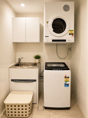 A compact laundry room with a washing machine, a dryer, a sink, and a small plant. - Modern Apartment Near Stadium & Shops (Lidcombe)