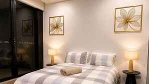 A hotel room with a checkered bedspread, two bedside lamps, and wall art. - Modern Apartment Near Stadium & Shops (Lidcombe)