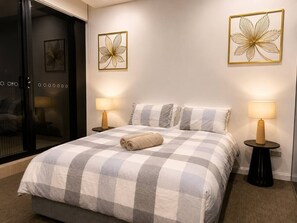 A hotel room with a checkered bedspread, two bedside lamps, and wall art. - Modern Apartment Near Stadium & Shops (Lidcombe)