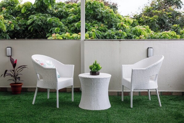 A patio area with two white wicker chairs and a small table, surrounded by artificial green grass and a wall with a potted plant.