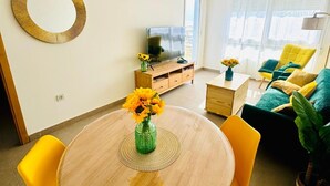 Una sala de estar moderna con una mesa redonda de madera, sillas amarillas, un sofá con cojines verdes, un televisor y un ventilador de techo.