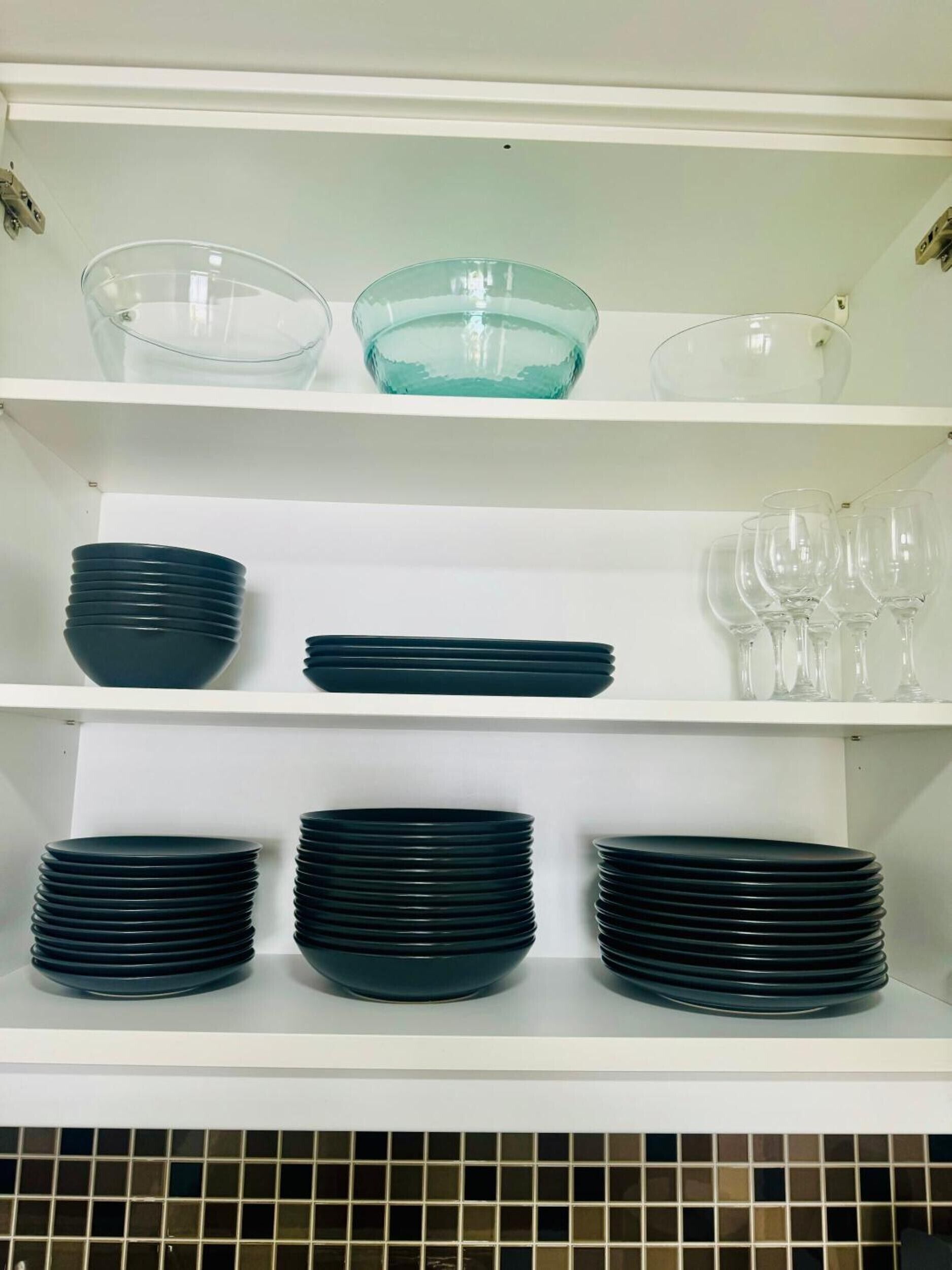 Un estante de cocina con platos negros apilados, un bol de vidrio azul verdoso y copas de vino transparentes.