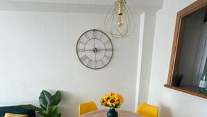 Comedor con una mesa redonda, sillas amarillas, un jarrón con girasoles, un reloj de pared y una lámpara colgante.