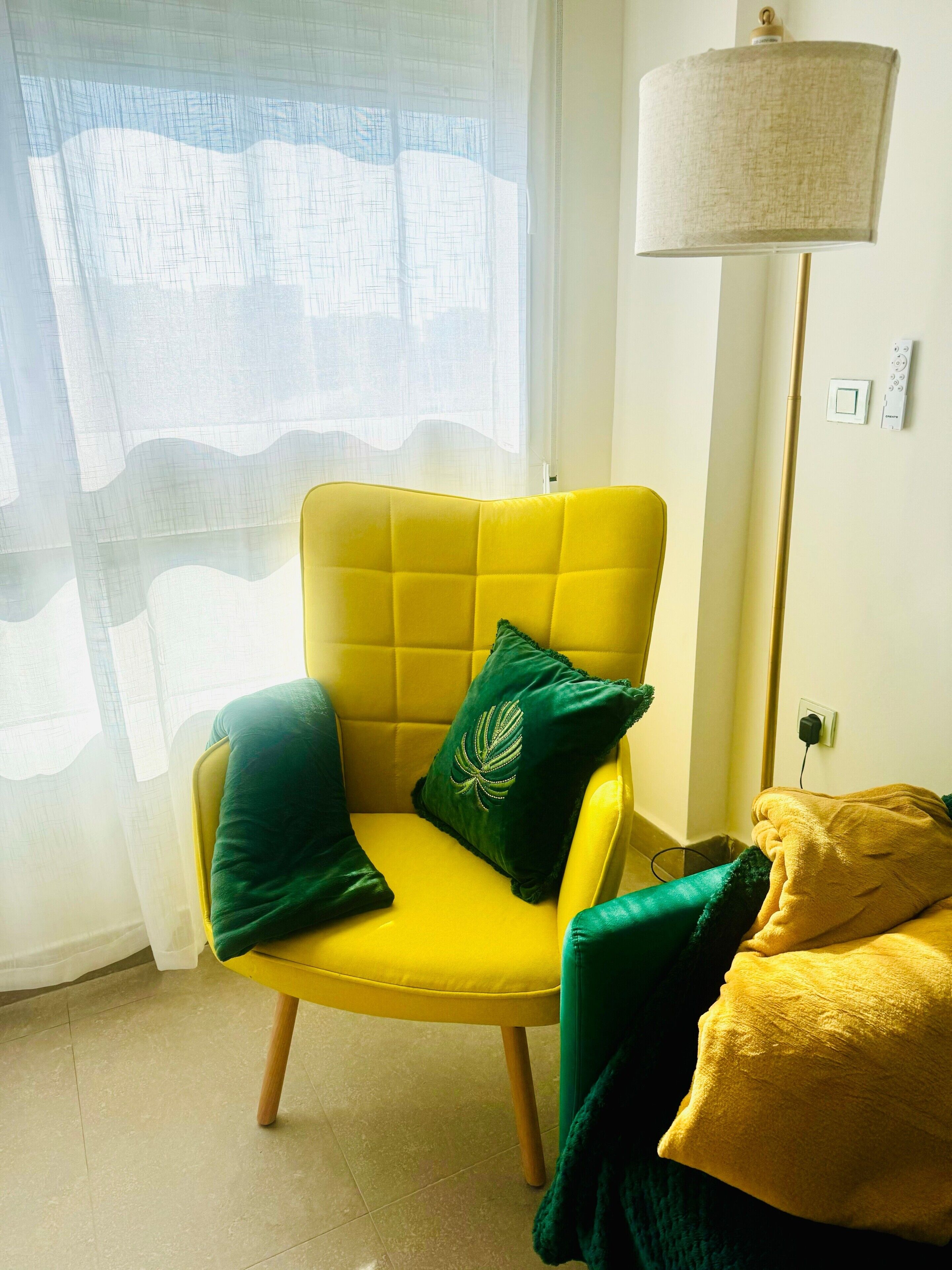Ein gelber Sessel mit grünen Kissen, eine Stehlampe und ein Fenster mit durchscheinenden Vorhängen.