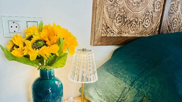 Una mesita de noche de madera con una lámpara, un jarrón con girasoles y una cama con una colcha verde.