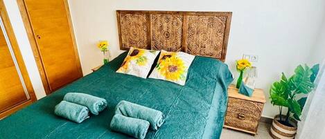 Una cama bien tendida con una colcha verde y dos toallas enrolladas, un cabecero de madera con intrincados grabados, una mesita de noche con una planta y un pequeño cofre de madera.
