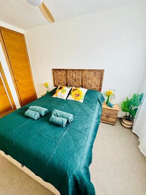 Una cama bien tendida con una colcha verde y dos toallas enrolladas, un cabecero de madera con intrincados grabados, una mesita de noche con una planta y un pequeño cofre de madera.