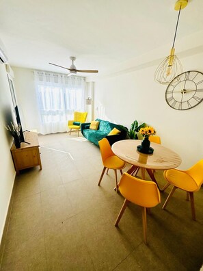 A modern living room with a dining area, a ceiling fan, a large window with curtains, and a wall clock. - Apartment 'Los Girasoles De Veneziola' with Sea View, Wi-Fi and Air Conditioning (La Manga del Mar Menor)
