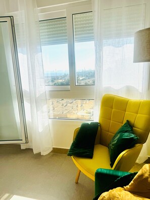 A yellow armchair with a green pillow, a window with blinds, and a view of a cityscape.