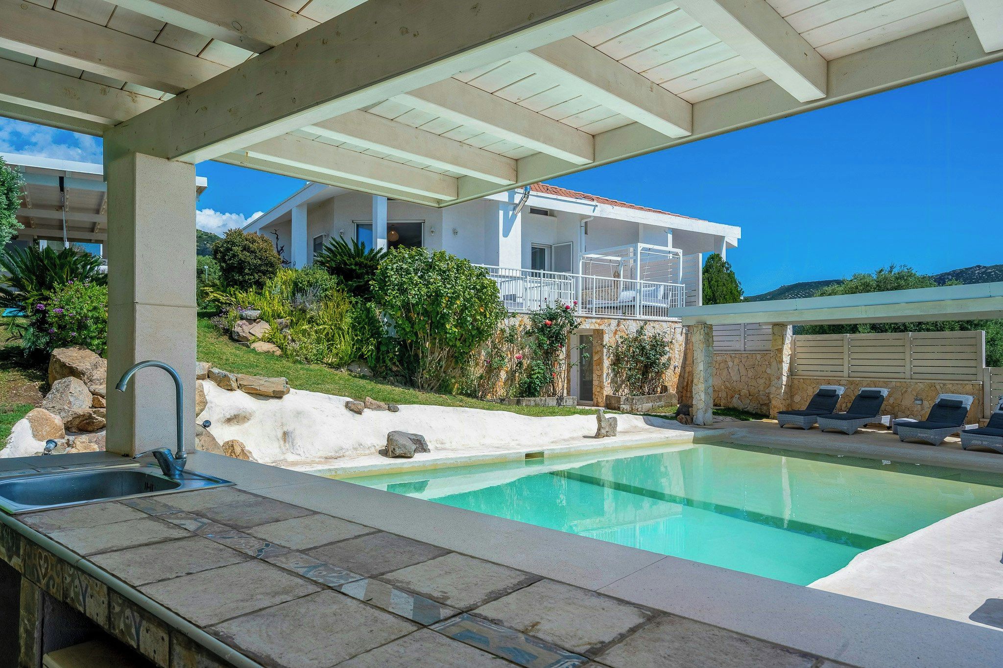 A swimming pool with a stone deck, a covered patio area, and a building with a balcony.