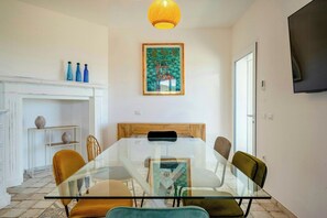 Een eetgedeelte met een glazen tafel en stoelen, een open haard met decoratieve flessen, en een schilderij boven de schoorsteenmantel.