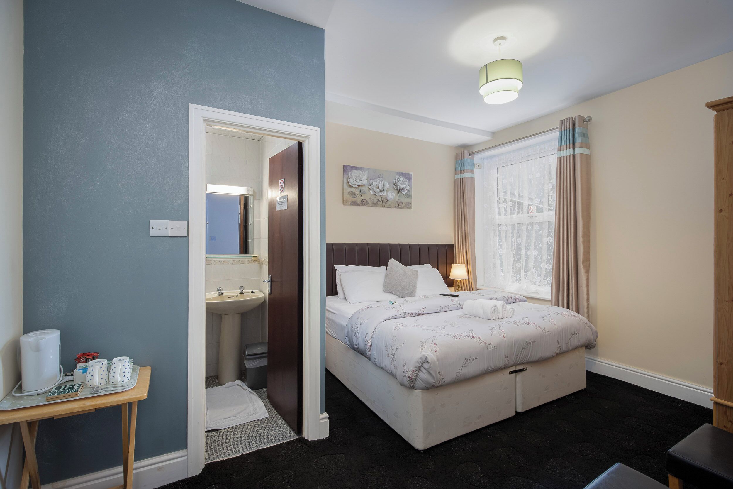 A hotel room with a bed, a bedside table with a lamp, a window with curtains, and a bathroom visible through an open door.