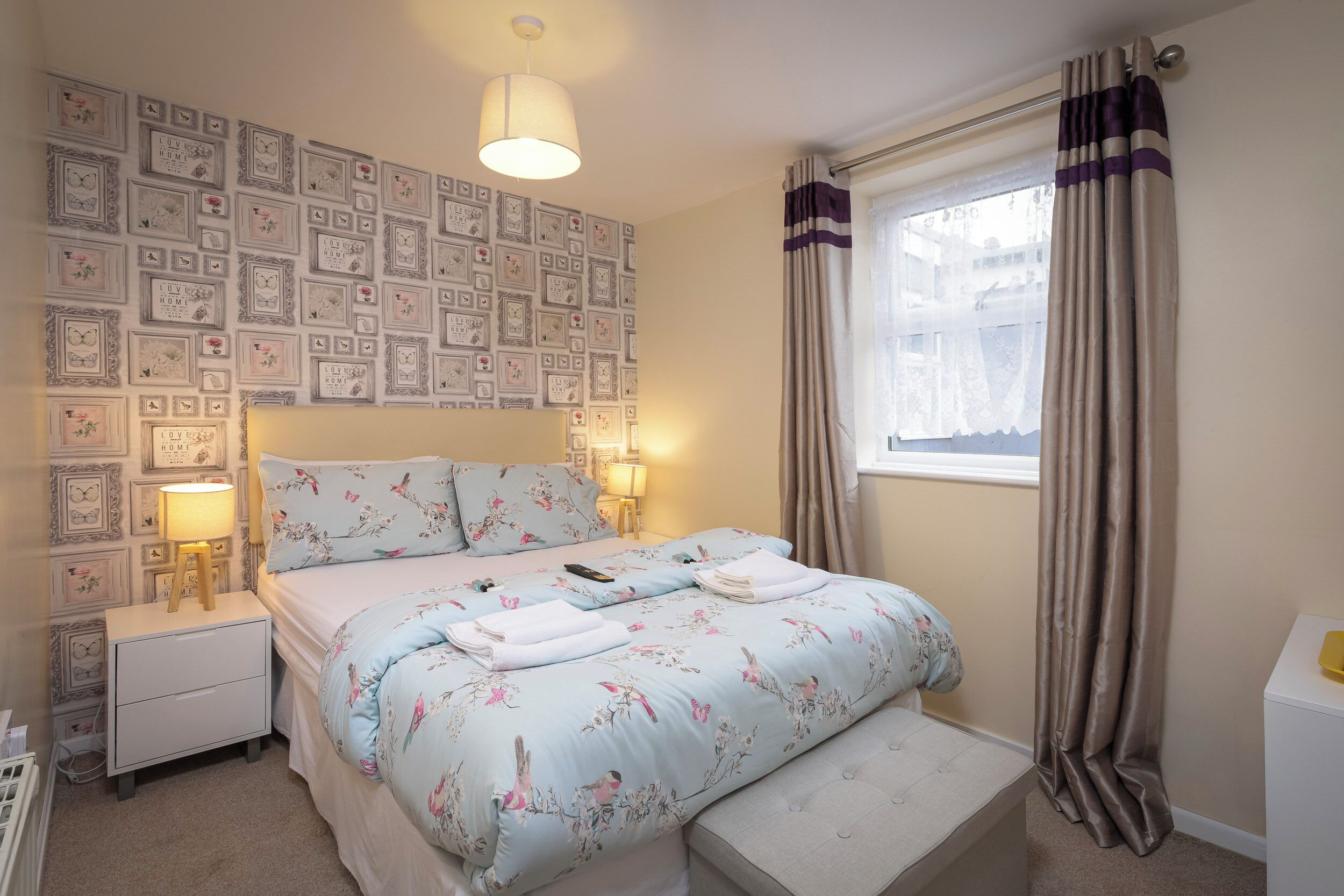 A bedroom with a bed, bedside tables, a lamp, a window with curtains, and a wall decorated with framed pictures.