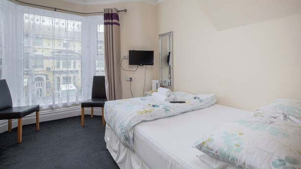 A hotel room with a bed, a chair, a television, and a window with curtains. - Devon Guest House (Blackpool)