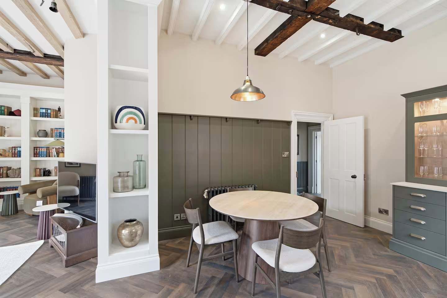 A dining area with a round table, chairs, a hanging light fixture, and a bookshelf with various items.
