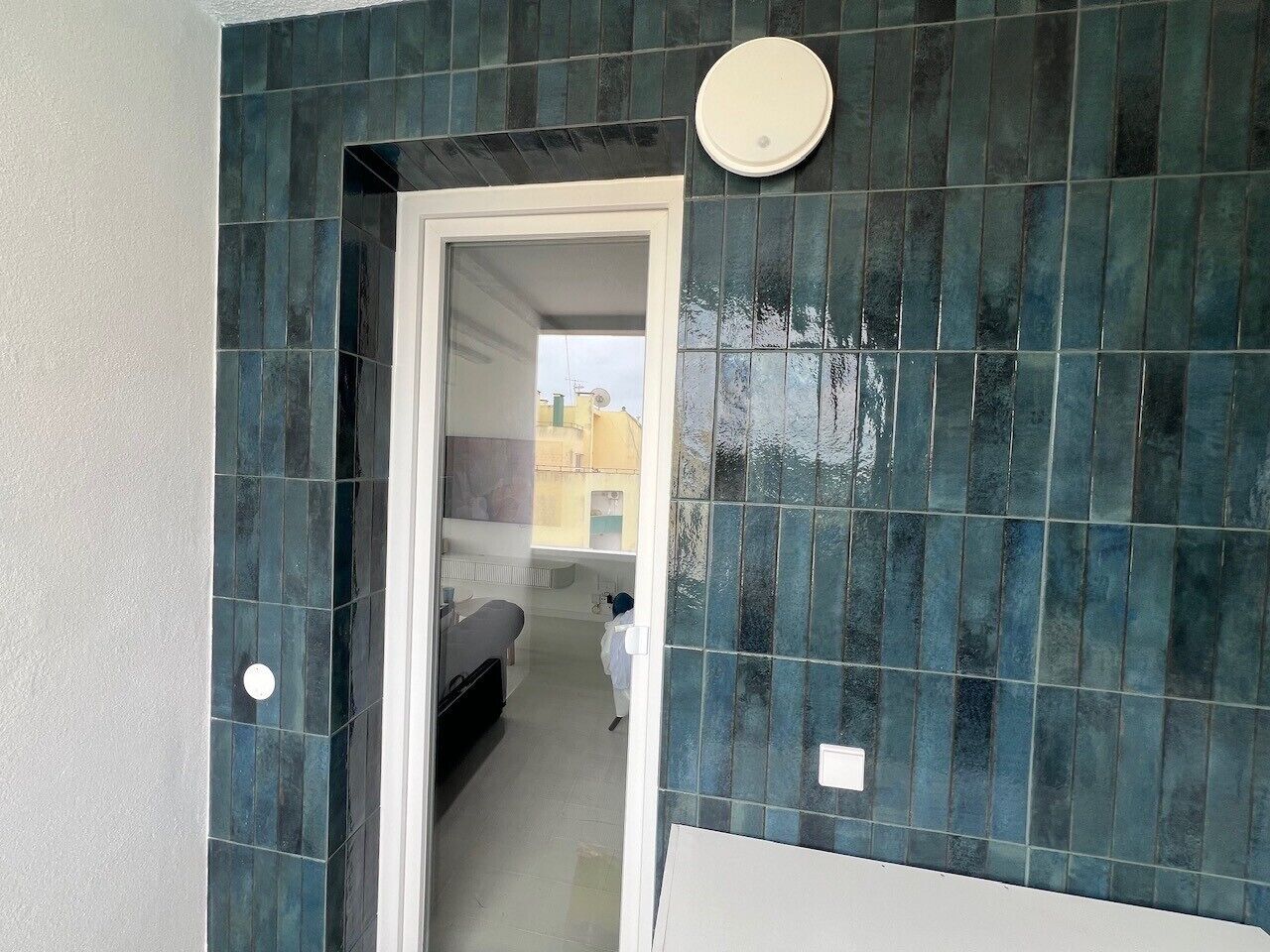 A bathroom with dark blue tiles, a white door, and a visible hallway leading to a room with a bed.