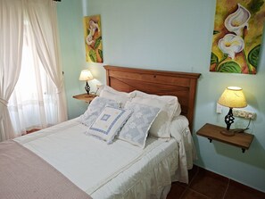 A neatly made bed with a patterned blanket and pillows, two bedside lamps, a wooden headboard, and a wall with floral paintings. - Casa Luciíta: Cozy, with a fireplace, patio, and BBQ. Near Marbella (Ojén)