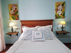 Un lit avec une literie blanche et des coussins à motifs, flanqué de deux tables de chevet avec des lampes, le tout adossé à un mur orné de deux tableaux représentant des motifs floraux.