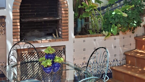 Une terrasse extérieure avec un four en briques, une table en verre avec des chaises et un mur recouvert de carreaux où sont disposées des plantes.