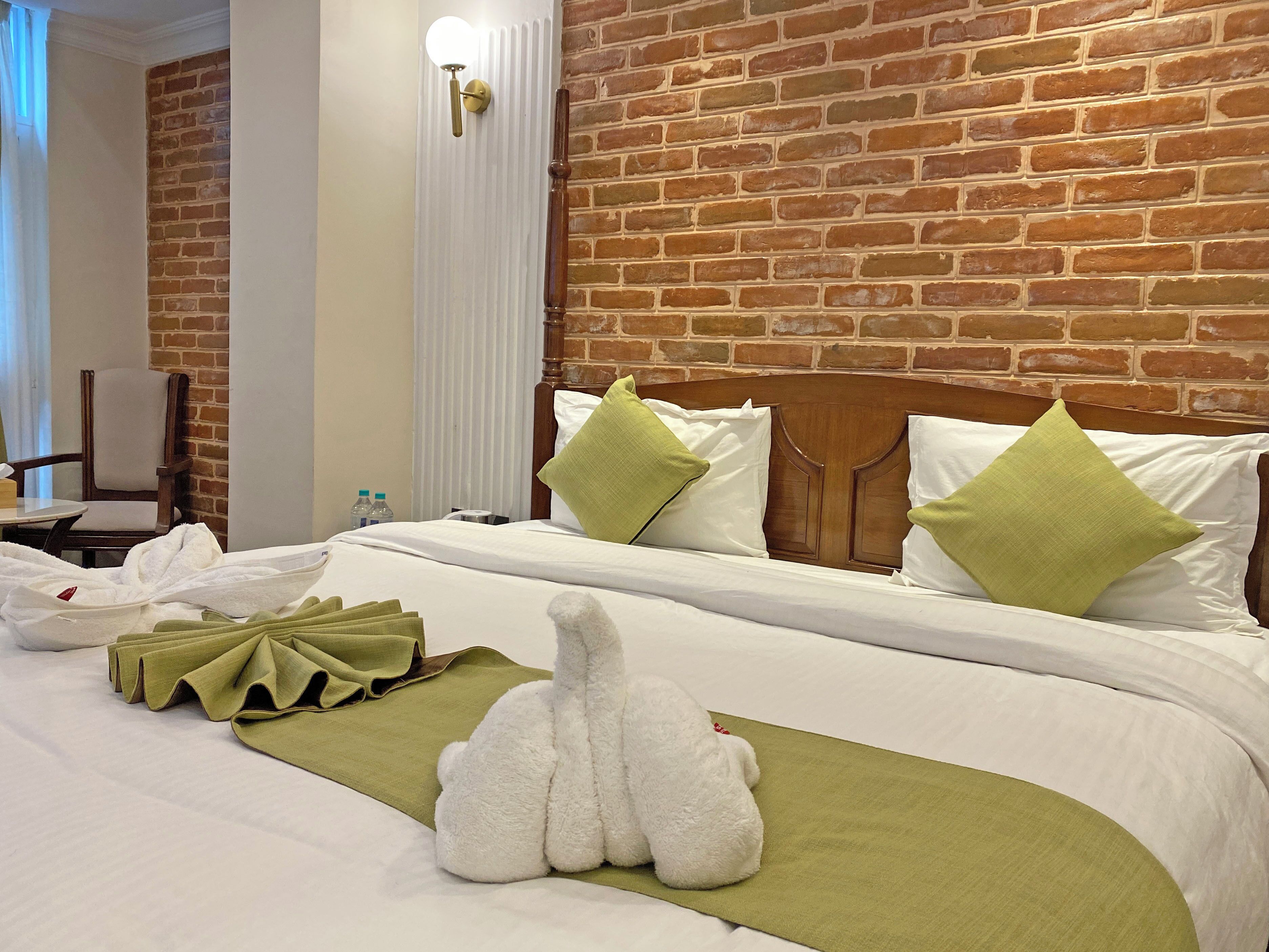 Een hotelkamer met een bakstenen muur, een houten hoofdeinde en een bed met witte lakens en groene kussens.