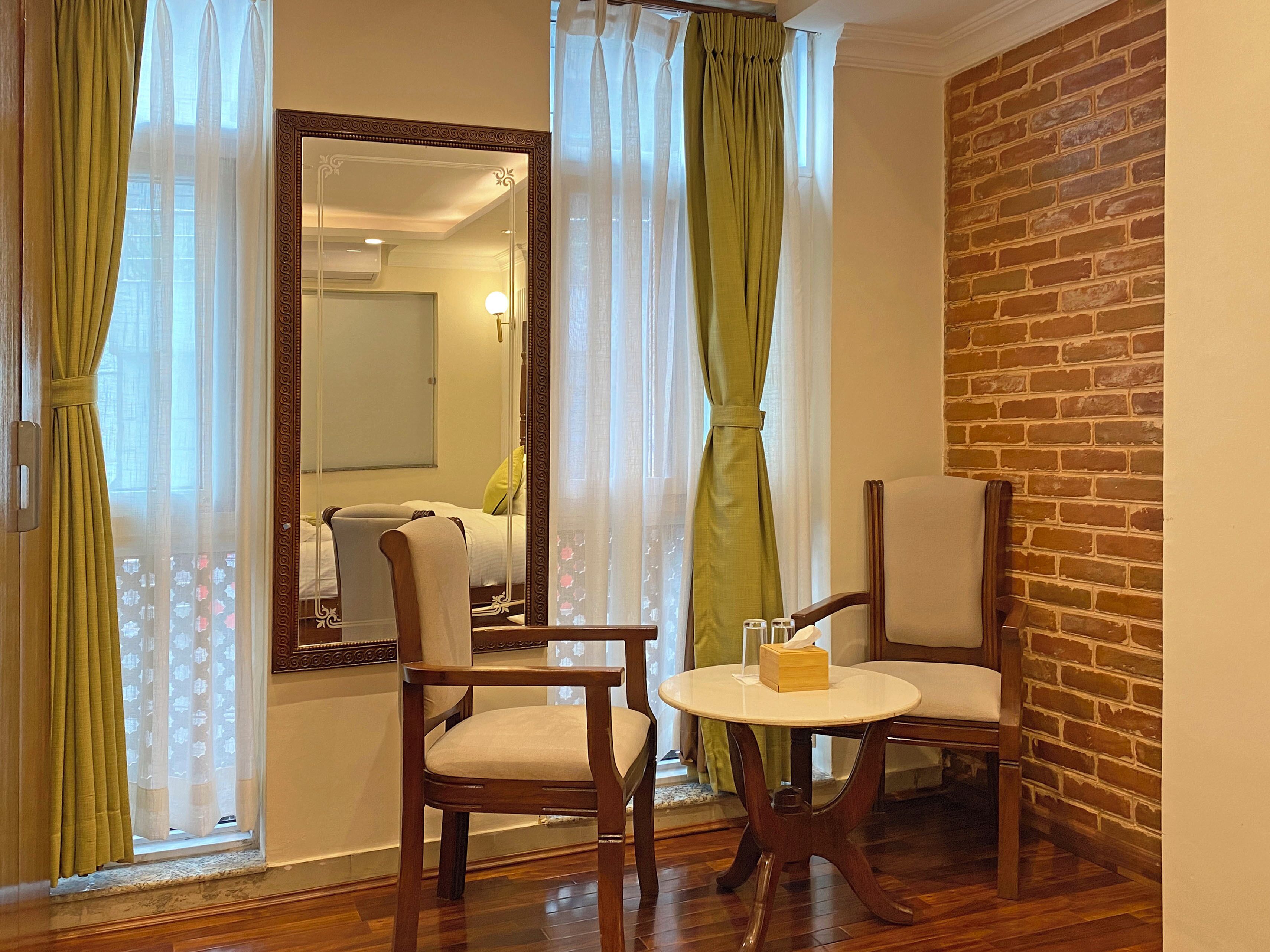 Una habitación con pared de ladrillo, suelo de madera, cama con ropa blanca, dos sillas de madera, una mesa redonda con una caja de pañuelos y un espejo con marco decorativo.