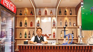 Een bar met schappen vol drankflessen, een Coca-Cola koelkast en een shisha op de toonbank.