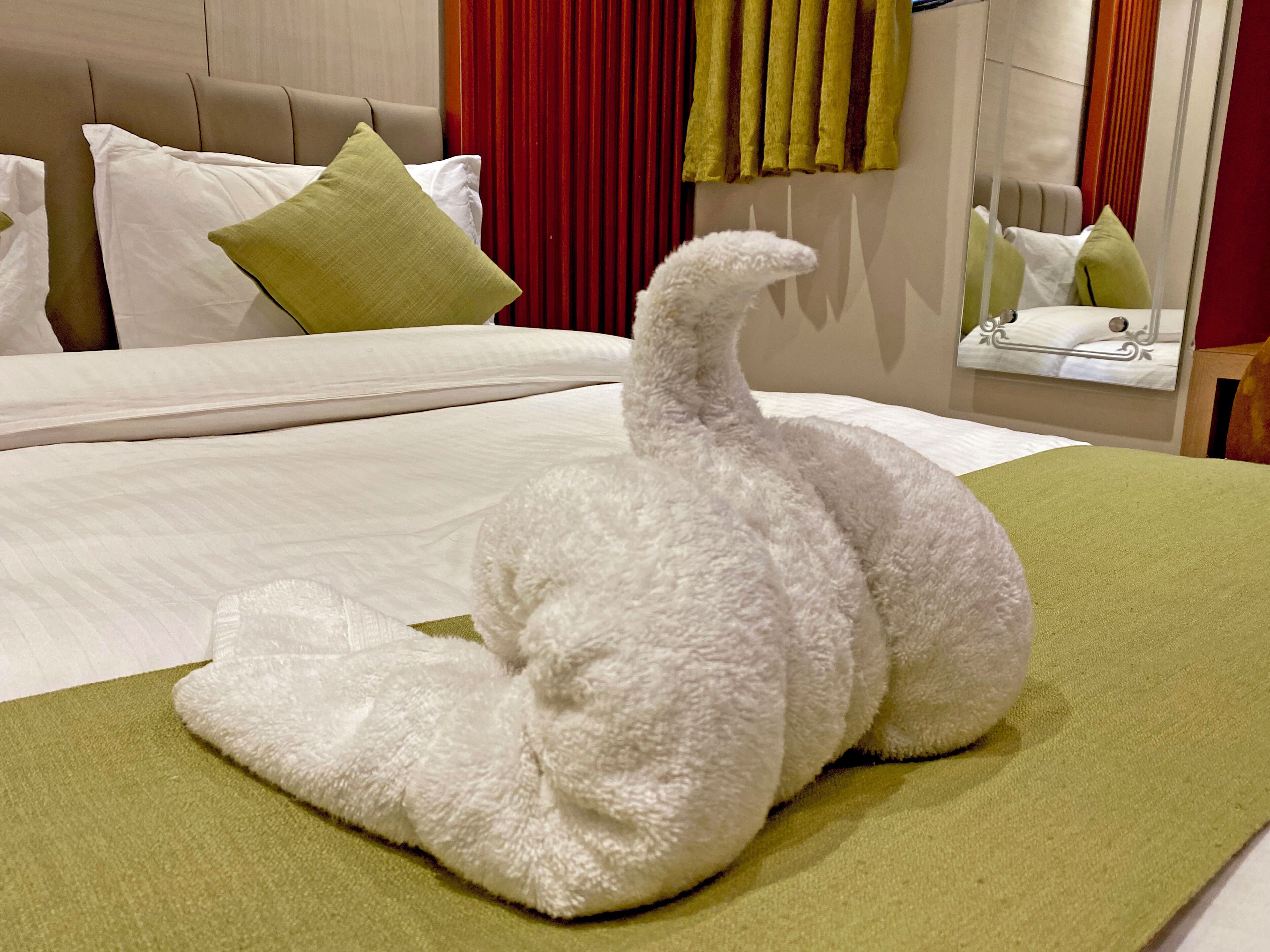 Una cama bien hecha con una almohada blanca, una almohada verde y un objeto blanco mullido que parece un cisne sobre un edredón verde.